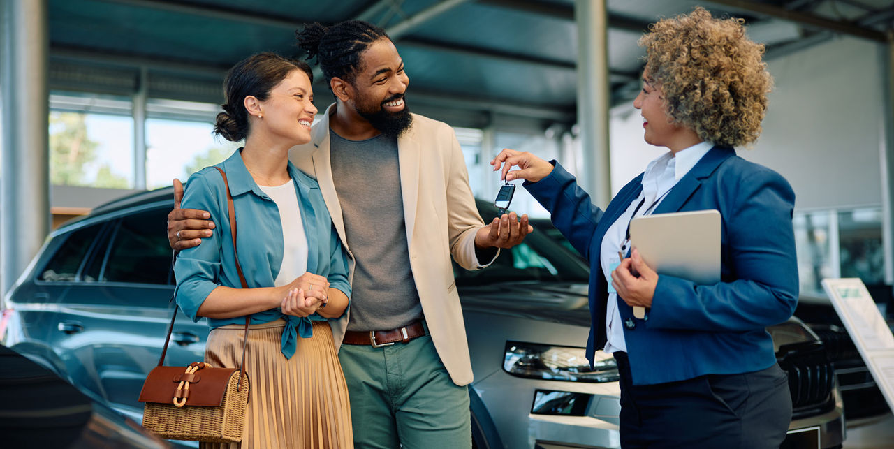 Happy multiracial couple receiving keys of their new car from saleswoman in showroom.